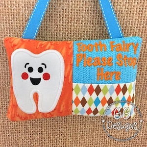 May include: A tooth fairy pillow with a smiling white tooth on an orange background. The pillow is made of fabric and has a blue ribbon for hanging. The text "Tooth Fairy Please Stop Here" is embroidered on the pillow in orange thread.