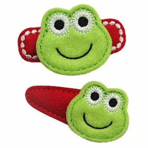 May include: Two green felt frog hair clips with red felt backing. The frogs have white embroidered eyes and a smile.