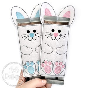 May include: Two felt bunny-shaped treat bags, one with blue ears and paw prints and the other with pink ears and paw prints. The bags are designed to hold a chocolate bar.