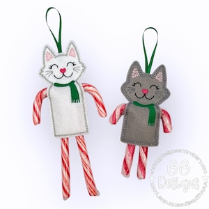 Puede incluir: Dos adornos de gato de fieltro con piernas de bastón de caramelo. El gato blanco tiene una bufanda verde y mejillas rosadas. El gato gris tiene una bufanda verde y mejillas rosadas. El texto "GG Designs" es visible en la esquina inferior derecha.