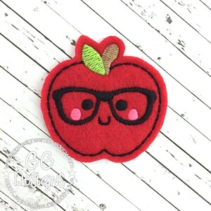 May include: Red felt apple with a green stem and brown leaf, wearing black-rimmed glasses and a smiling face. The apple has pink cheeks.