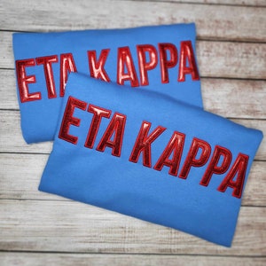 Op de afbeelding: Twee blauwe sweatshirts met de tekst "Eta Kappa" in rode glitterletters.