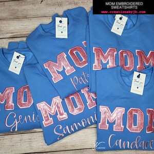 May include: Four blue sweatshirts with the word "MOM" in pink glitter lettering and a personalised name below. The sweatshirts are stacked on top of each other.