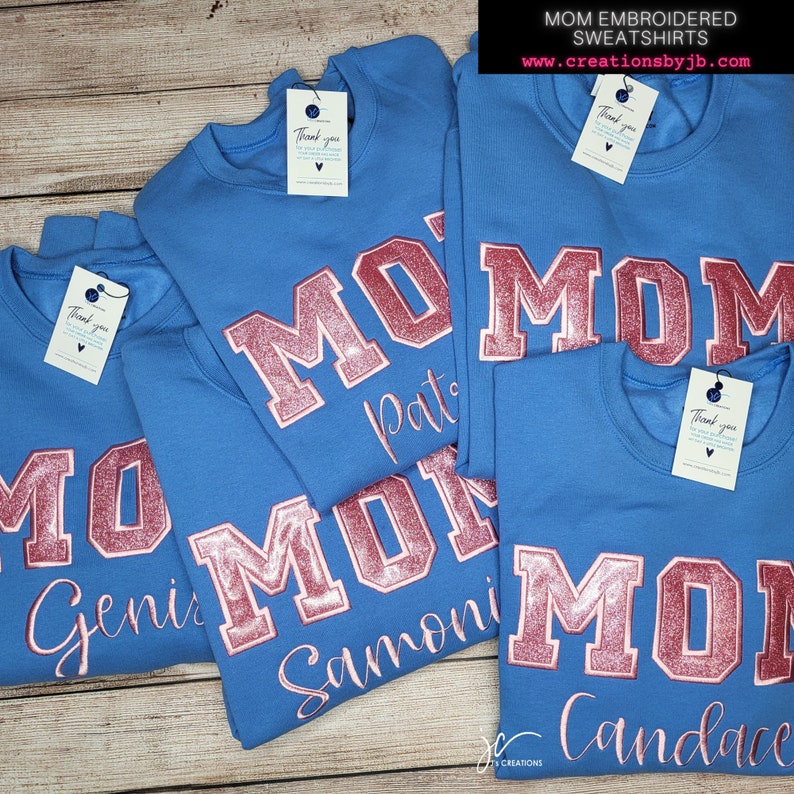 May include: Four blue sweatshirts with the word "MOM" in pink glitter lettering and a personalised name below. The sweatshirts are stacked on top of each other.