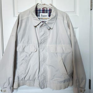 May include: A beige zip-up jacket featuring a classic collar, two front flap pockets, and buttoned cuffs. The interior is lined with a multicolored plaid fabric. A label at the inner neckline displays the text London Fog with a small building icon.
