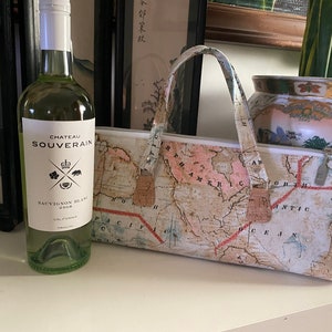 Op de afbeelding: Een fles witte wijn met een etiket waarop "Chateau Souverain Sauvignon Blanc 2018 Californië" staat, naast een beige draagtas met een kaartprint. De kaartprint toont een vintage kaart van Noord-Amerika met de woorden "Noord-Amerika" en "Atlantische Oceaan" zichtbaar.