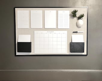 Könnte beinhalten: Ein schwarzer, gerahmter Korktafel mit einem Kalender, Notizblöcken und einer kleinen Vase mit einer Pflanze. Der Kalender ist mit "November" beschriftet.