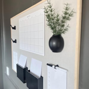 May include: A corkboard with a white calendar labeled "March", a black vase with a green plant, two black mail holders with white paper, a white clipboard with a black pen, and a black and white patterned background.