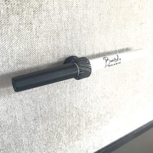 May include: A black marker with a white cap is held in place by a black metal holder. The marker has the text "Board's Balance" written on the cap.