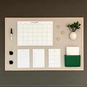 May include: A large rectangular wall-mounted magnetic board with a light wood frame. It features a white November calendar, three blank white note sheets, a black marker, three wooden magnets, a small white vase with a plant, and a green pocket.