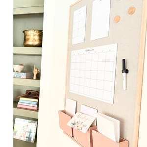 May include: A rectangular wall-mounted bulletin board with a light wood frame and a beige fabric surface. It features a white February calendar, two blank white sheets, two round wooden magnets, a black marker, and three light pink storage pockets.