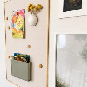 May include: A rectangular beige bulletin board with a light wood frame, featuring small round wooden magnets, a white ceramic wall vase containing yellow dried flowers, and a sage green rectangular pocket holding colorful paper art prints.