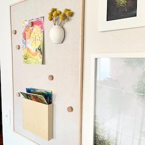 May include: A light beige cork board with a wooden frame, featuring a small vase with yellow dried flowers, a colorful drawing, and a wooden magazine holder with colorful magazines. The cork board is mounted on a white wall.