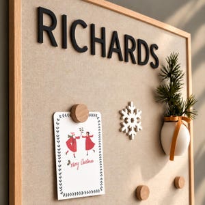 May include: A square wooden-framed bulletin board with a beige fabric surface. The black text "RICHARDS" is mounted at the top. A white card with a red-clad figure illustration and "Merry Christmas" text is held by a round wooden magnet.
