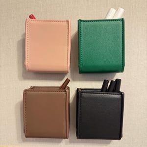 May include: Four small, square pen holders in a grid. The pen holders are in shades of pink, green, brown, and black. Some contain pens or pencils. The pen holders are made of a smooth material and have a stitched border.
