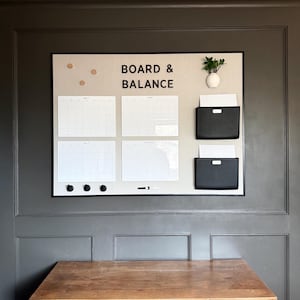 May include: A rectangular wall-mounted organization board with a light beige fabric surface, featuring the text BOARD & BALANCE. It includes four white monthly calendar grids, two black document pockets, three round wooden pins, and a small white vase.