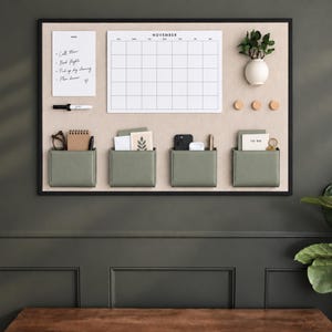 May include: A rectangular wall-mounted organization board with a beige fabric surface and black frame. It features a white November calendar, a notes pad, four sage green storage pockets, three wooden knobs, and a small white vase with greenery.