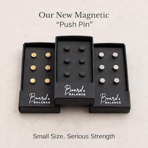 May include: Three black rectangular boxes labeled Board & Balance, each containing six magnetic push pins. The left box holds gold-toned pins, the center box holds black pins, and the right box holds silver-toned pins. Text reads Our New Magnetic Push Pin.