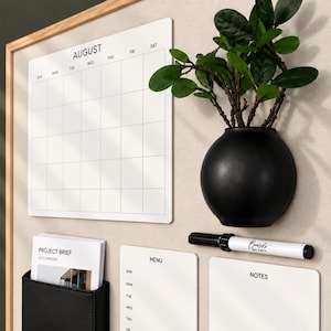 May include: A wall-mounted organization board with a light wood frame featuring a white August calendar, a black spherical wall planter with a green plant, a black pocket holding a Project Brief, a menu board, a notes board, and a black marker.