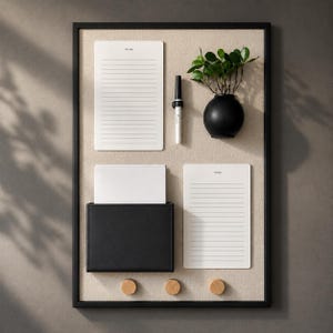 May include: A rectangular wall-mounted organizer board with a black frame and beige fabric backing. It features two white lined to-do notepads, a black marker, a small black vase with green leaves, a black pocket, and three round wooden hooks.