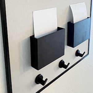 May include: A black wall-mounted organizer with two rectangular pockets holding white paper and three black hooks below. The organizer is framed in black and mounted on a light-colored background.