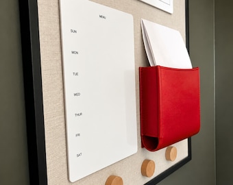May include: A wall-mounted organizer board with a black frame and beige fabric backing. It features a white weekly menu board with days of the week, a red pocket holding white paper, and three round light-colored wooden pegs at the bottom.