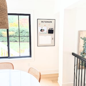May include: A rectangular magnetic wall board with a black frame, featuring the name PETERSON in black text at the top. The board includes a black storage pocket, a white monthly calendar grid, and a small black marker attached to the surface.