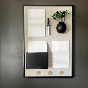 May include: A black framed corkboard with three white to-do lists, a black pen, a black container, three wooden push pins, and a small potted plant with green leaves in a black vase.