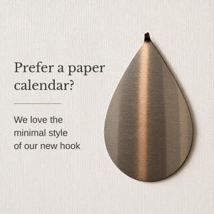 May include: A teardrop-shaped metal hook with a brushed bronze finish. The text on the left reads: Prefer a paper calendar? We love the minimal style of our new hook. The hook has a pointed top and a rounded base, featuring a metallic texture.