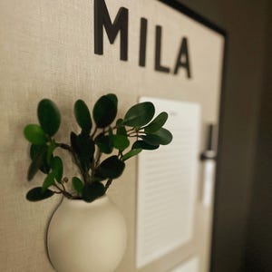 May include: A wall-mounted beige fabric board featuring the black text MILA at the top. A white spherical wall planter holds a green leafy plant. A white lined notepad is attached to the board, along with a black pen held in a clip on the side.