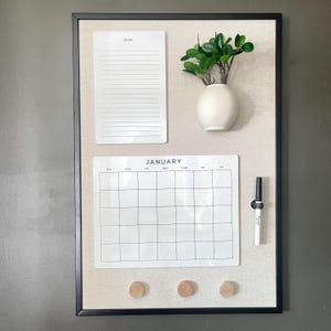May include: A rectangular wall-mounted organizer board with a black frame and beige fabric backing. It features a white lined to-do list, a white January calendar, a small white vase with green leaves, a black marker, and three wooden knobs.