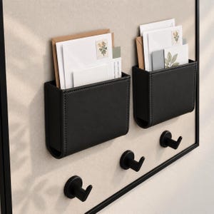 May include: A wall-mounted organizer featuring two black rectangular pockets with visible stitching and three matching black hooks. The pockets contain white envelopes with floral stamps. The entire unit is framed in a thin black border.