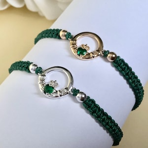 May include: Two braided dark green cord bracelets featuring circular metal charms. One charm is gold-toned and the other is silver-toned, both displaying a central green heart-shaped stone topped with a small crown and flanked by two hands.