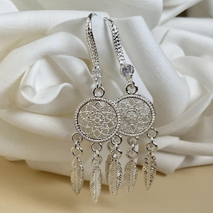 May include: Silver dangle earrings with a circular filigree design and feather charms. The earrings have a decorative pattern and are suspended from a curved ear wire with a small, clear gemstone accent.