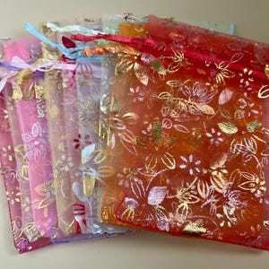 May include: A collection of colourful organza gift bags. The bags are in shades of pink, orange, yellow, and blue. They feature a gold floral design and matching drawstring ribbons. The bags are made of sheer material.