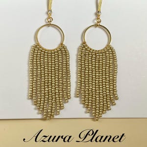 May include: A pair of gold-toned dangle earrings featuring a circular hoop frame with multiple strands of small metallic beads hanging in a fringe pattern. The earrings include a hook-style closure and are displayed on a white card with Azura Planet text.