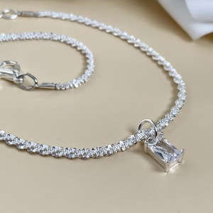 May include: A silver anklet with a textured chain. It has a rectangular, clear gemstone pendant. The clasp and connecting hardware are silver. The jewellery is displayed on a neutral, light-coloured surface.