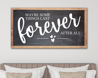 May include: A black chalkboard sign with a wood frame, hanging above a bed with white bedding. The sign reads "Maybe some things last forever after all" with two small hearts.