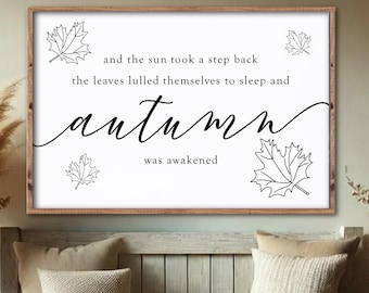 May include: A framed wall art piece with the word "autumn" in cursive script. The art features a wooden frame, white background, and black text. The text reads "and the sun took a step back, the leaves lulled themselves to sleep and autumn was awakened."