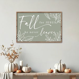 May include: A wooden sign with a sage green background and white lettering that reads "Fall for Jesus he never leaves". The sign is decorated with white leaf outlines. The sign is hanging on a white wall above a wooden bench with white pumpkins and orange pumpkins.