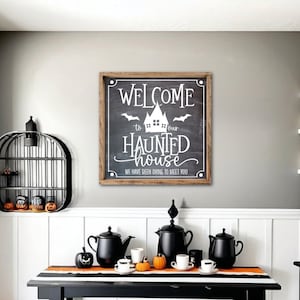 May include: A black and white striped table runner with black teapots and cups on a black table. A sign with the text "Welcome to our Haunted House. We have been dying to meet you." is on the wall behind the table.