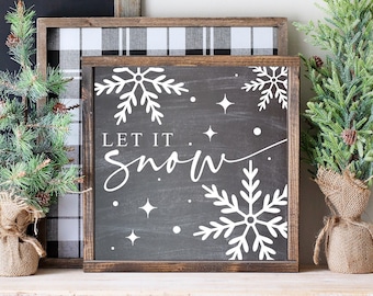 May include: A black chalkboard sign with white lettering that reads "Let it snow". The sign is decorated with white snowflakes and stars. The sign is sitting on a white shelf with two small artificial pine trees on either side.