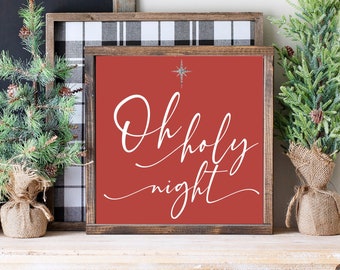 May include: A framed sign with the words "Oh holy night" in white script on a red background. A silver star is above the text. The sign is surrounded by a wooden frame and sits on a white shelf with two small Christmas trees.