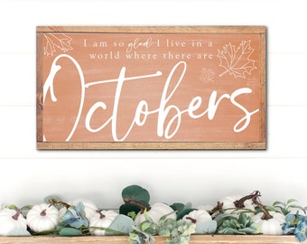 May include: A rectangular wooden sign with the word "Octobers" in white script, above a wooden trough filled with white pumpkins and greenery. The sign has the text "I am so glad I live in a world where there are Octobers".