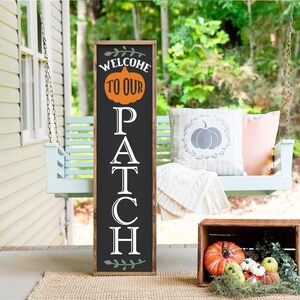 May include: A wooden sign with a black background and white lettering that reads "Welcome to our Patch". The sign is decorated with a green vine and a pumpkin. The sign is hanging on a porch swing.