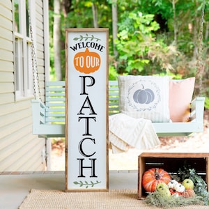 May include: A wooden sign with the words "Welcome to our Patch" in black lettering. The sign is decorated with a green leaf design and a pumpkin with a white stem. The sign is leaning against a white porch swing.