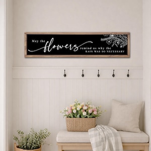May include: A rectangular wooden sign with the text "May the flowers remind us why the rain was so necessary" in white script on a black background. The sign is framed in light brown and features a floral design.