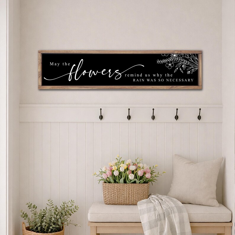 May include: A rectangular wooden sign with the text "May the flowers remind us why the rain was so necessary" in white script on a black background. The sign is framed in light brown and features a floral design.