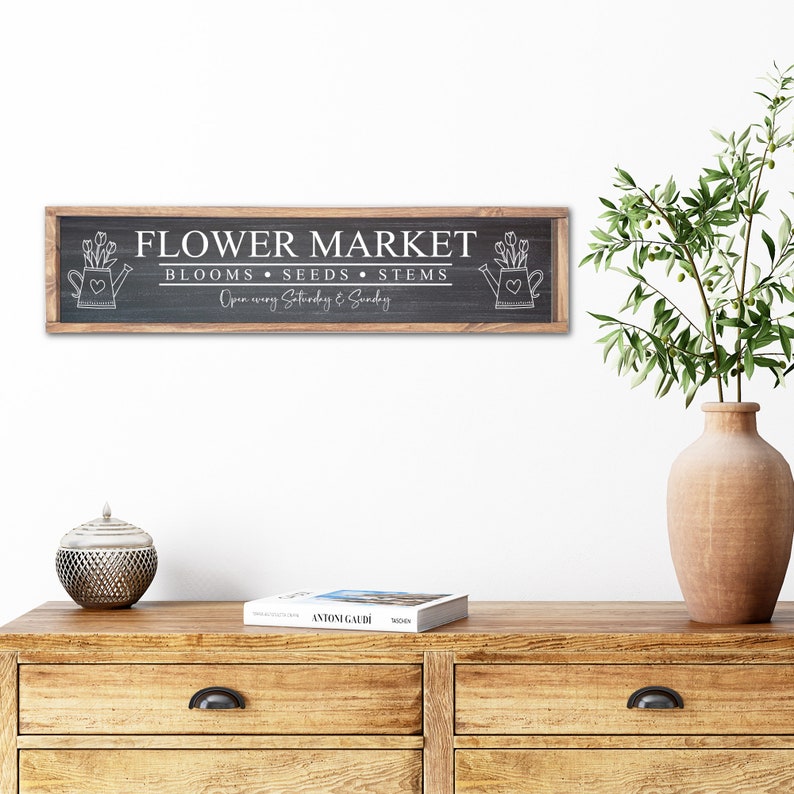 以下が含まれることがあります： 木製のフレームが付いた白黒の看板で、「Flower Market」と「Blooms, Seeds, Stems」と「Open every Saturday & Sunday」の文字が書かれています。看板の両側には、花が咲いたジョウロの絵が描かれています。
