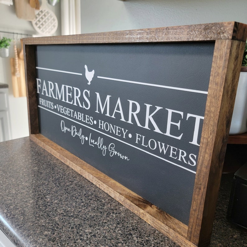 K&ouml;nnte beinhalten: Ein dunkelgraues Holzschild mit wei&szlig;em Text, der "Farmers Market" lautet, mit einer wei&szlig;en Silhouette eines Huhns &uuml;ber dem Text. Das Schild sagt auch "Fruits, Vegetables, Honey, Flowers" und "Open Daily, Locally Grown".